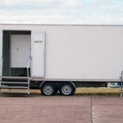 A white portable restroom trailer labeled "GENTS" with stairs leading to the entrance, parked outdoors on a paved surface, with grass, barriers, and tents visible in the background.